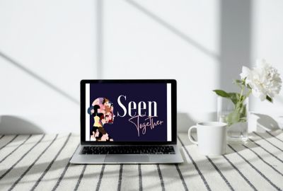 Mockup of a laptop on a white wooden table next to a white mug and white flowers in a clear glass vase. On the laptop is a graphic with a silhouette of a woman's head filled with smaller illustrations of different women. To the right is Seen Together in bold white font against a navy blue background.